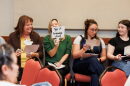 Four women sit next to each other, holding pieces of paper. One woman is smiling as another holds up a card that reads "type of language used". 