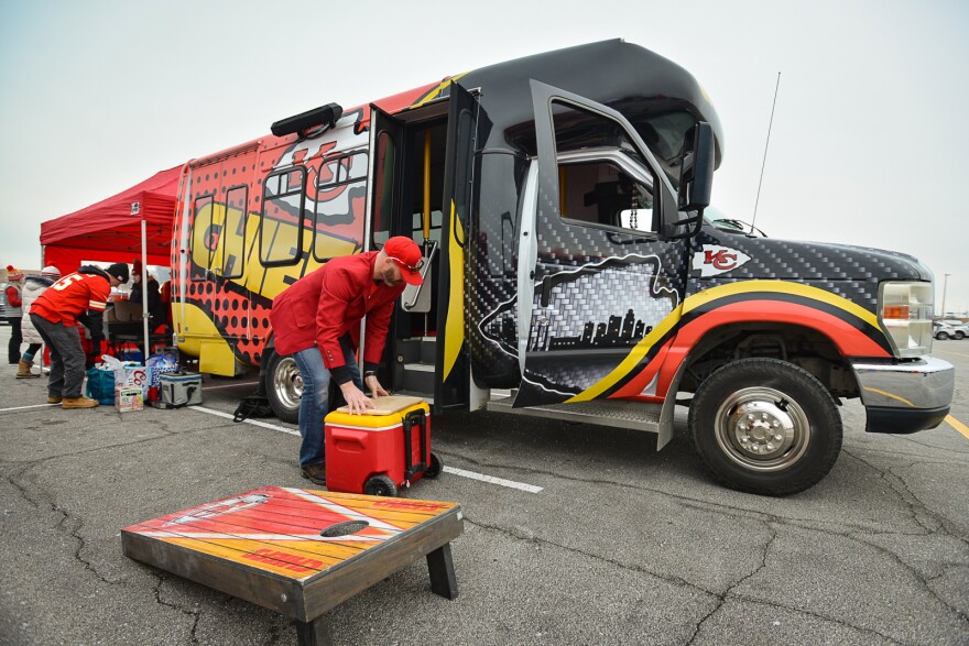 A man wearing a red jacket and red ball cap sets down a yellow and red ice chest from a large, colorful van that has Kansas City Chiefs wording and logos printed on it.