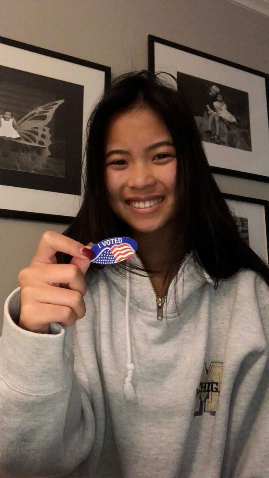 A young woman wearing a gray sweatshirt takes a photo of herself holding an 'I Voted' sticker.