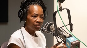 A woman inside a radio studio sits behind a microphone talking. She is gesturing with both hands.