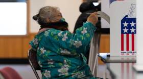 A woman in a flowery parka and a mamsk puts her ballot in a box