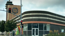 A mid-century brick building with a curved, black brickwork entryway and a brick clock tower sit at an intersection. Small, white lettering above the front door simply reads, "The Drugstore."