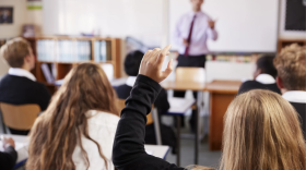 Un estudiante levanta la mano en un aula, junto a otros niños y con el profesor al frente.