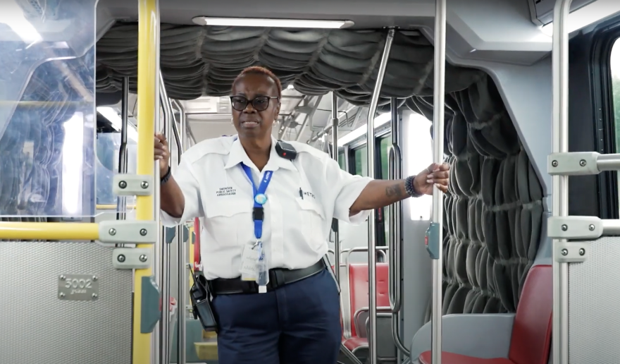 A public safety ambassador in the official uniform of white shirt and navy blue pants stands inside a Capital Metro bus, holding the support bars for stability while the vehicle moves.