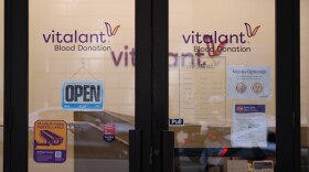 Exterior shot of two glass doors of a blood donation center. An open sign is hanging on the inside of the left door. Hours of operation are printed in white on the right door. On both doors, in purple and black text, "Vitalant" is printed. Inside, the same logo is visible on the wall, along with a table with plastic water bottles