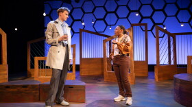 UGA students Jacob Arkin and Samara McKnight standing facing each other on a starkly dressed stage with brown, angled, wood and wire set pieces in front of a glowing blue-lit backdrop covered by the silhouette of a honeycomb pattern.