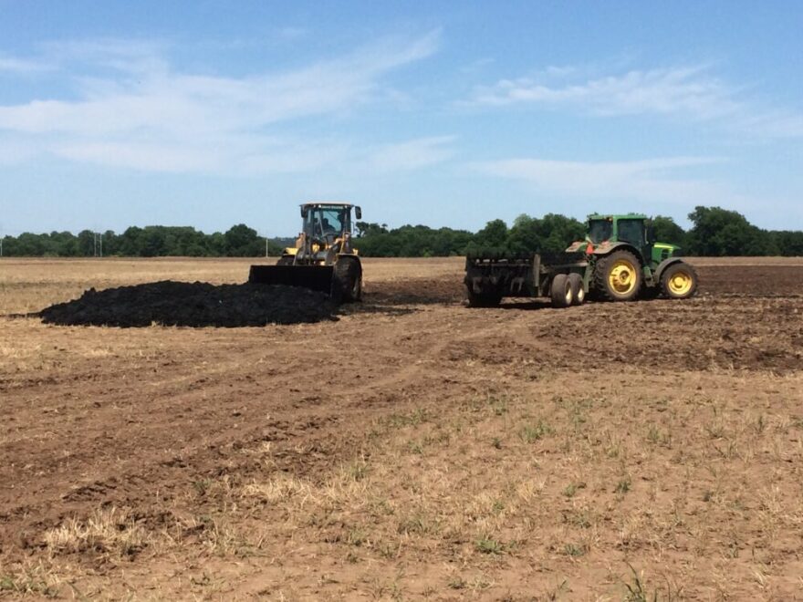 A photo of two tractors spreading a black-looking substance on dirt farm fields. The sky is blue and there are green trees at the edge of an open field