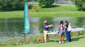 A long white folding table on the banks of White's Lake holds 4 white plastic tubs with different soil samples in them.  The instructor wears a blue golf shirt and khaki pants.  Two female students and an instructor stand listening to him.  One girl has long black braids with pink tips, blue jean cut off shorts and a sleeveless white tank top.  The girl next to her in blue jeans and a black t-shirt carries a white back pack.  A female teacher with blond hair is barely seen next to the girl with the back pack.  A red golden retriever runs along the shore near a First Green banner.  Lilly pads grow abundantly in the still lake. 