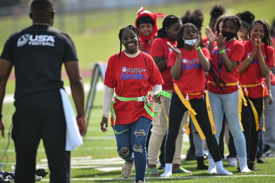 A man wearing a dark T-shirt and pants is seen from the back. In front of him, girls wearing red T-shirts that read "Play Football" are lined up on a football field. They are clapping and smiling as one girl runs toward the man.