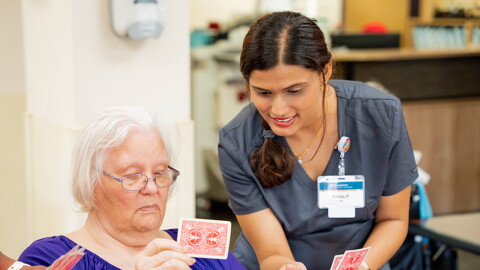 A nurse in scrubs leans over while holding playing cards next to a nursing home resident, also holding playing cards.