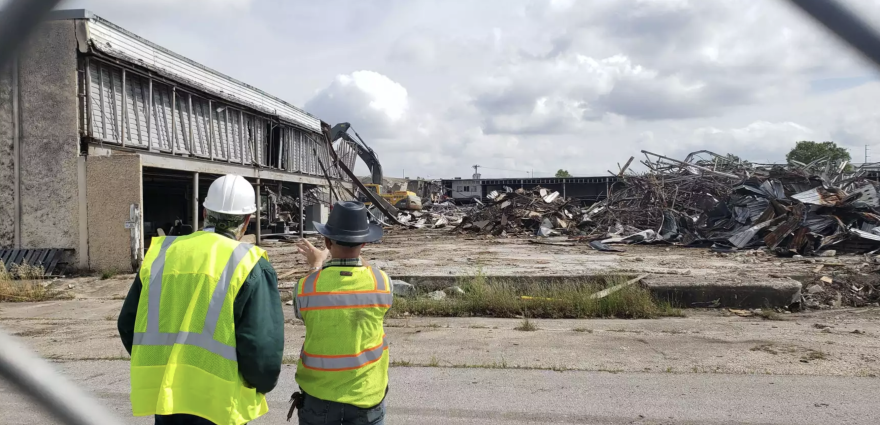 Two construction workers in high-visibility vests and hard hats observe the demolition of an old building. The site is filled with rubble and debris, with an excavator in the background actively tearing down the structure. The sky is overcast with scattered clouds.
