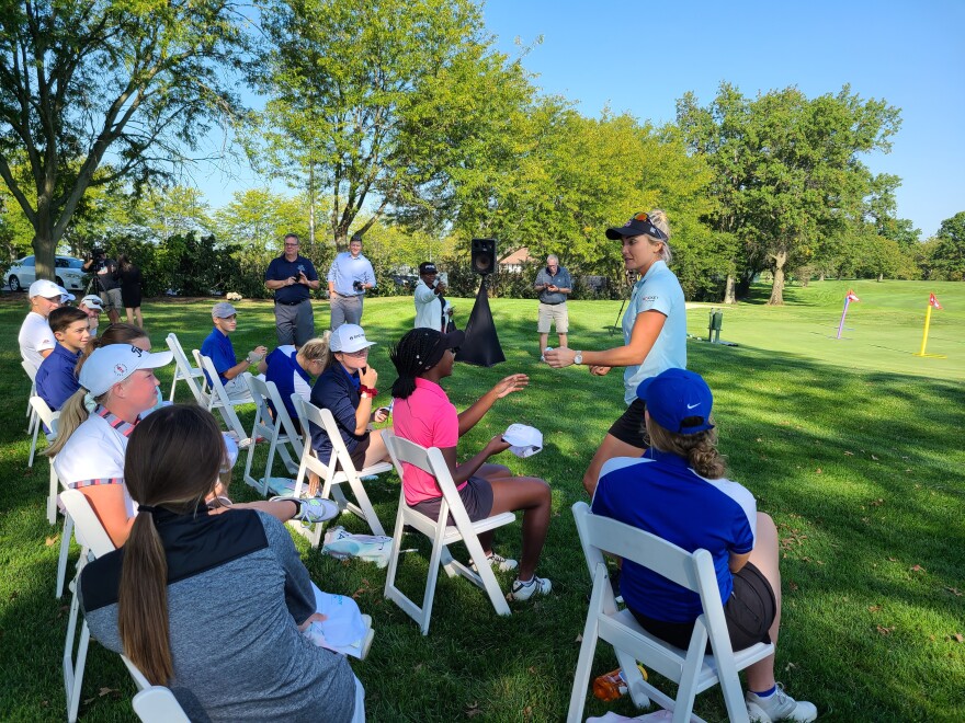 two rows of seated high school age girls on a golf course watch as Lexi Thompson signs autographs. She is handing a signed golf ball back to one of them. The s