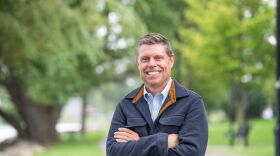 A smiling man with short brown hair standing with his arms crossed. He is wearing a blue collared button-down shirt under a blue fleece with a brown lining. 