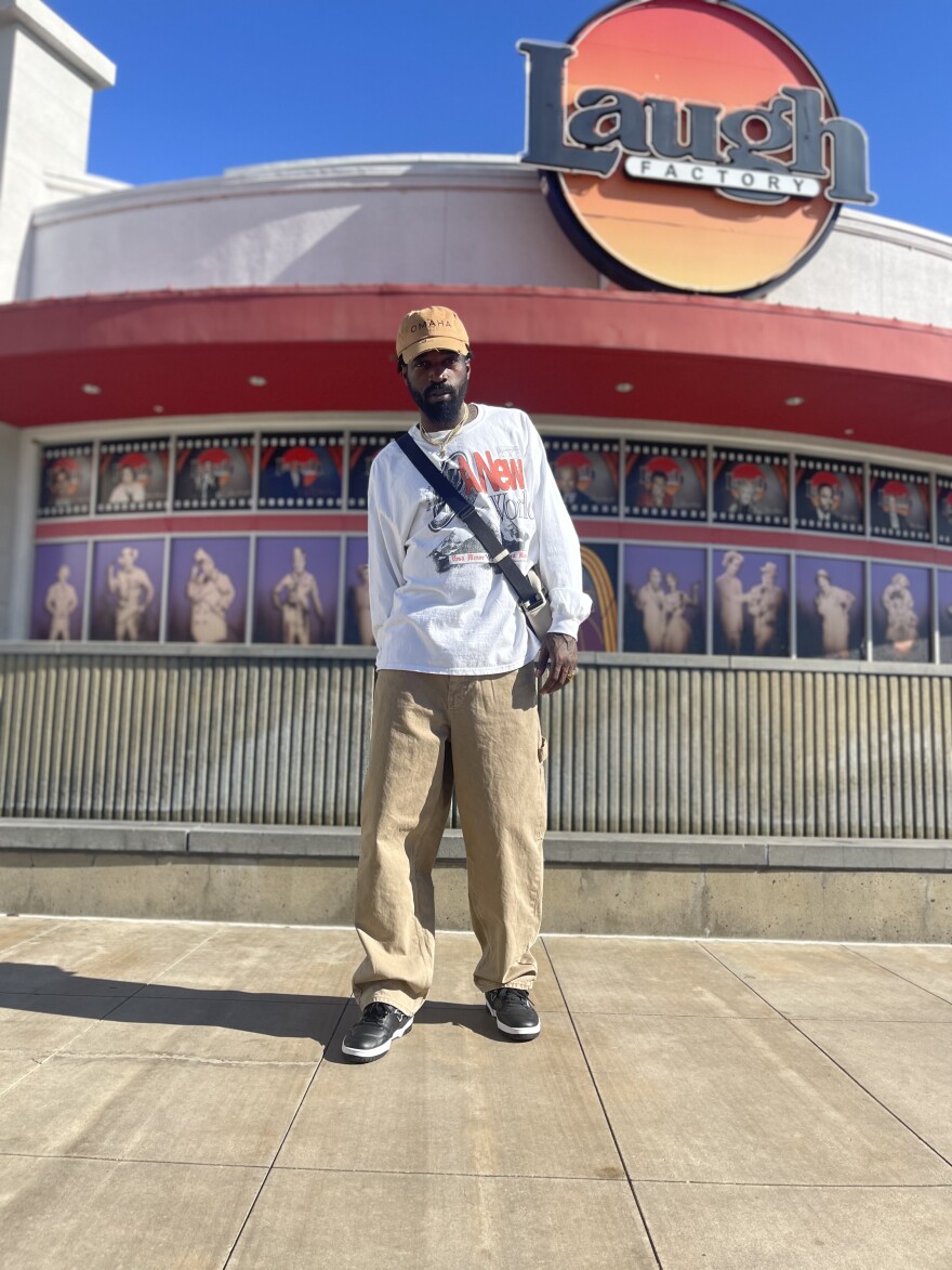 Marcey Yates is a Black man with a dark beard. In this photo, he's wearing a baseball cap, white hoodie and tan pants. He's posing in front of The Laugh Factory in San Diego.