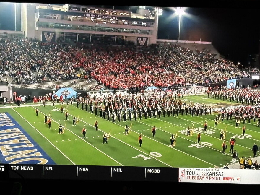 A college football stadium with a large marching band performing on the field. The stands are full of fans, many wearing red, and stadium lights illuminate the scene at night. A TV sports ticker runs along the bottom of the screen.