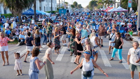 A large crowd fills a palm tree-lined street at an outdoor evening concert, with dozens of people dancing in the foreground and spectators seated in lawn chairs along the sidewalks under vendor tents in the background.