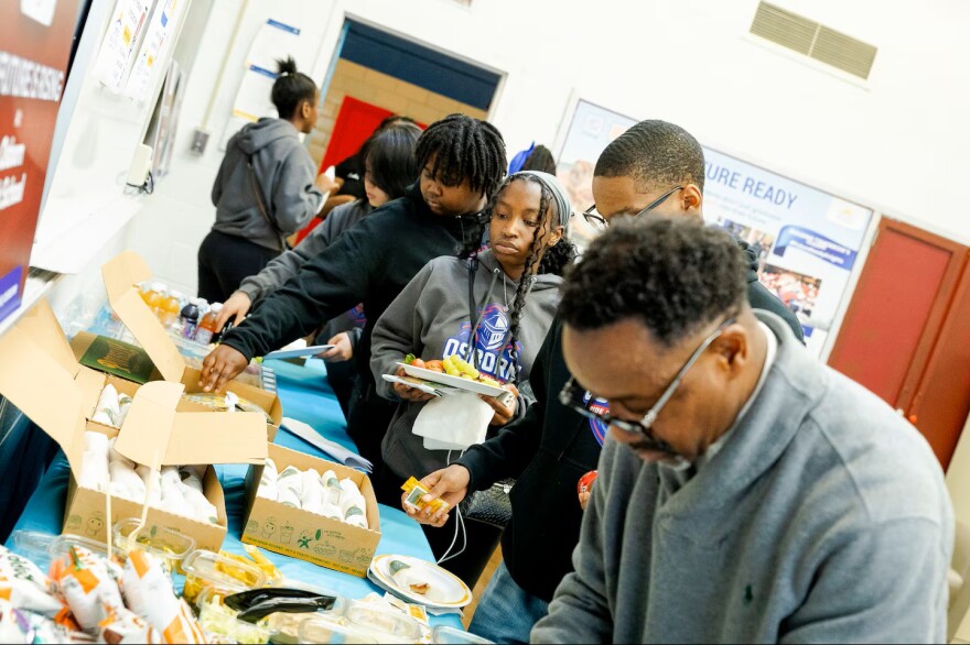A line of teenagers moves down a buffet table covered with blue tablecloths, picking up food options. The teen in the center wears a grey hoodie with an "Osborn" graphic logo and holds a white plate stacked with fresh fruit and food. In the foreground, an older Black man with glasses and a grey sweater leans forward over the table.