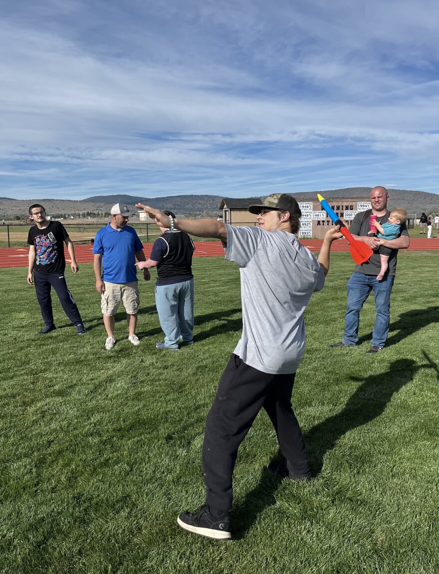 A teenaged boy in a gray t shirt and green hat prepares to throw a colorful plastic javelin. On the field behind him, a few people watch. 