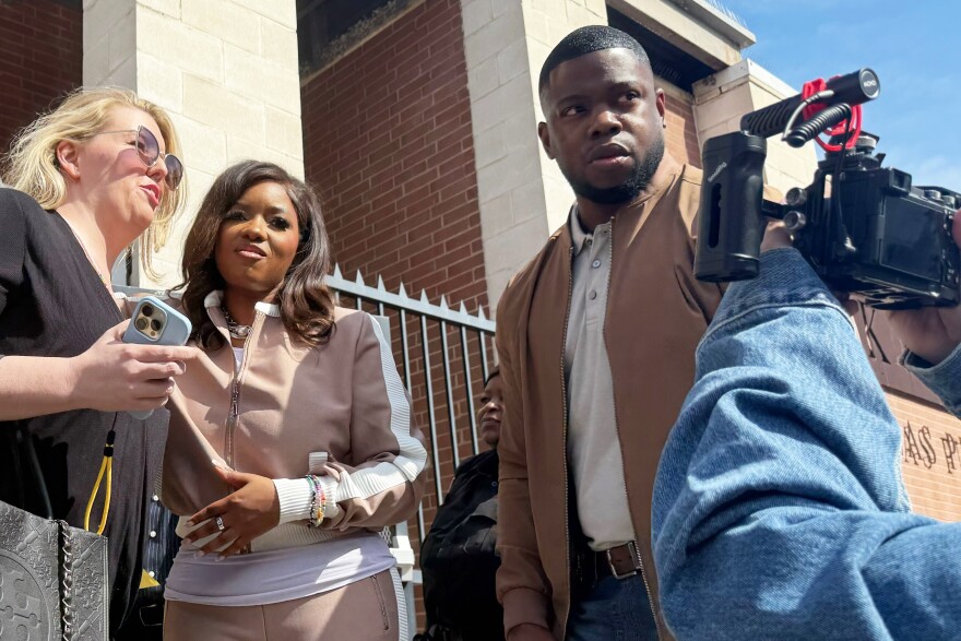 A blonde woman with sunglasses puts her arm around a woman in a brown track suit, next to a man in a brown jacket.