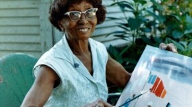 Clementine Hunter holding her artwork. Hunter worked as a house worker at Melrose Plantation, where she started out quilting and doing other crafts before an artist visiting the property gave her leftover paint and brushes. Body - 4