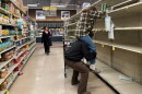 A man hoists a woman on his shoulders to reach water bottles on the top shelf of an otherwise empty set of shelves.