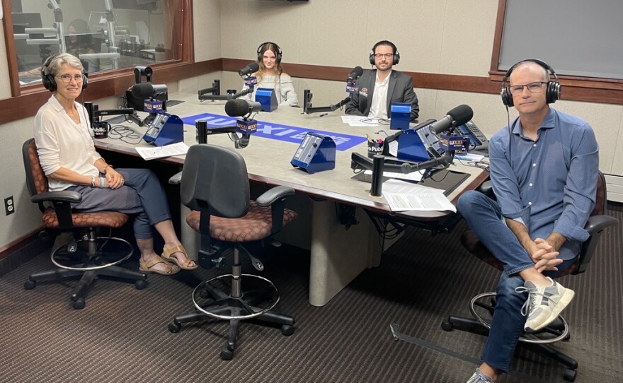 Four smiling people wearing headphones sit around a table in a radio talk studio: a woman front left has short grey hair and is wearing glasses, a white blouse, dark pants, and tan shoes; a woman back left has long light brown hair and is wearing a grey sweater; a man back center has short dark hair and a dark mustache and beard and is wearing a white button-down shirt under a grey suit coat; a man at right has very short dark hair and is wearing glasses, a blue button-down shirt, jeans, and grey and blue sneakers