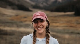 A woman wearing a light blue hoodie and a faded red ball cap, with her hair in two long, light-brown braids, smiles for a photo with mountains in the background