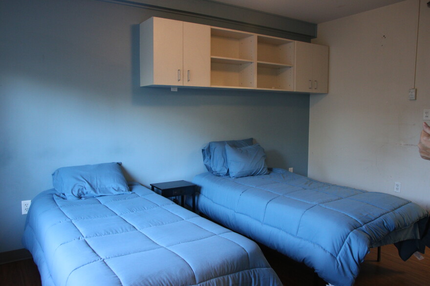 Two twin mattresses with blue comforters sit in a room with blue walls. Above the beds is a cream bookcase with storage space.