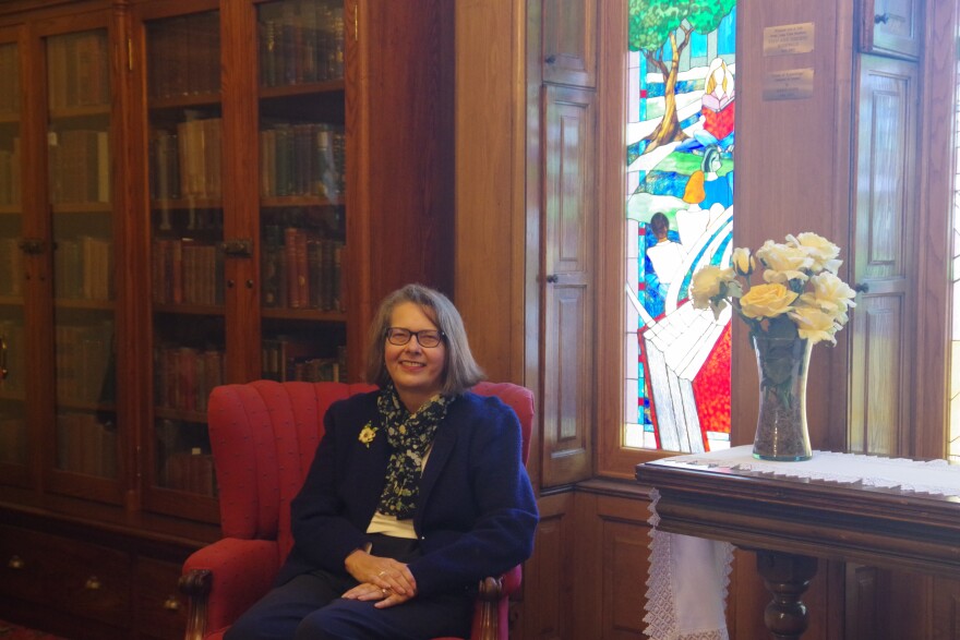 A woman wearing a blue suit jacket, white blouse and blue pants sits in a red chair, with a bouquet of yellow flowers standing on a table to the right. A long case of books stretches off on the left side of the image.  