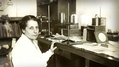Black-and-white photo of physicist Lise Meitner seated at a laboratory desk, surrounded by scientific instruments and papers, circa 1930.