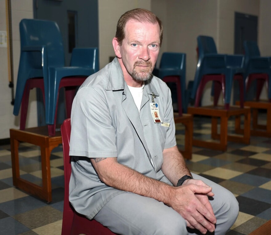 A man is sitting in a room surrounded by stacked, colorful chairs. He's wearing a gray prison jumpsuit and looking directly at the camera. 