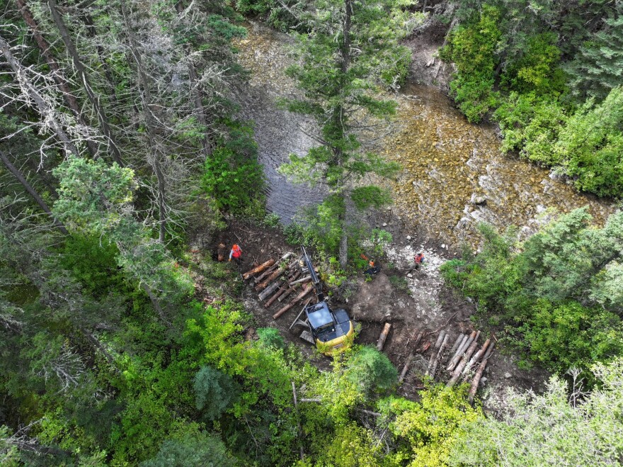 An aerial view of bank restoration on the Middle Fork Judith River. The photo shows what looks like a small feller buncher placing logs in a row perpendicular to the stream. Three workers in orange safety gear stand nearby.