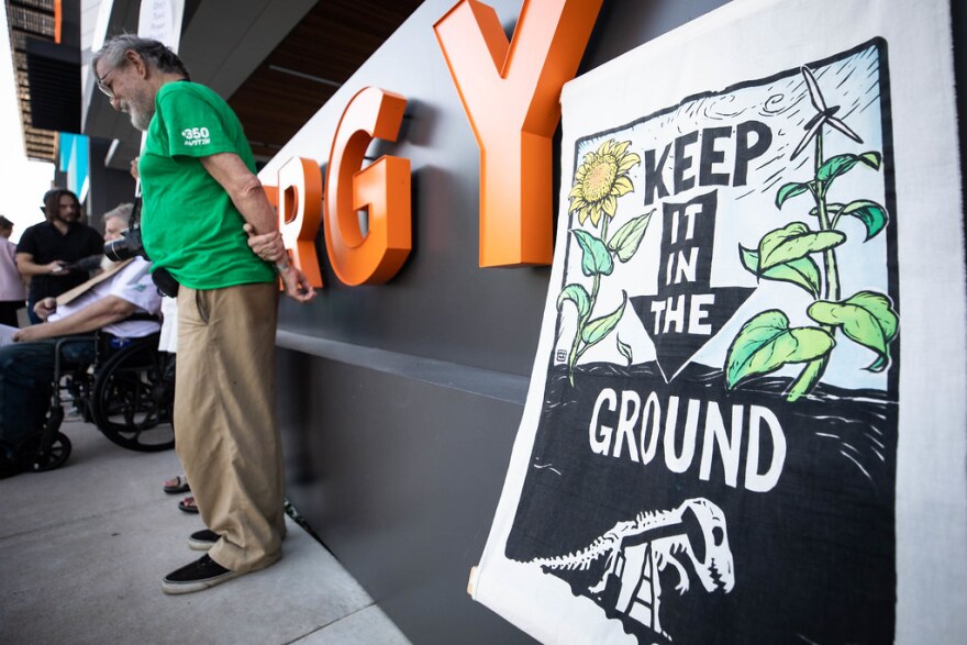 A man stands near a sign that says "Keep it in the ground" that features a pumpjack inside a dinosaur skeleton