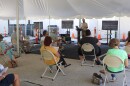 People sitting in chairs, facing Hudson, who is at a podium speaking with a screen behind him, all over a large tent the people are under.