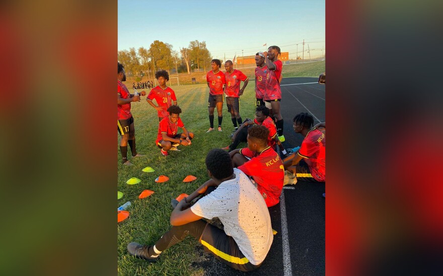 A groups of male soccer players stand and sit on the edge of a track near a soccer field. They appear to be listening to one man talking to the group.