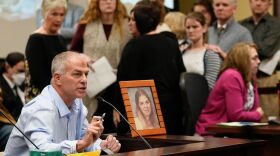 In a room crowded with people, a white man with graying hair sits at a table, speaking into a microphone. Beside him is a framed picture of a woman.