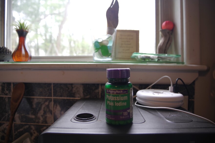 A green bottle labeled "Potassium plus Iodide" sits in front of a windowsill. Outside the window, trees can be seen. Small trinkets sit on the windowsill.