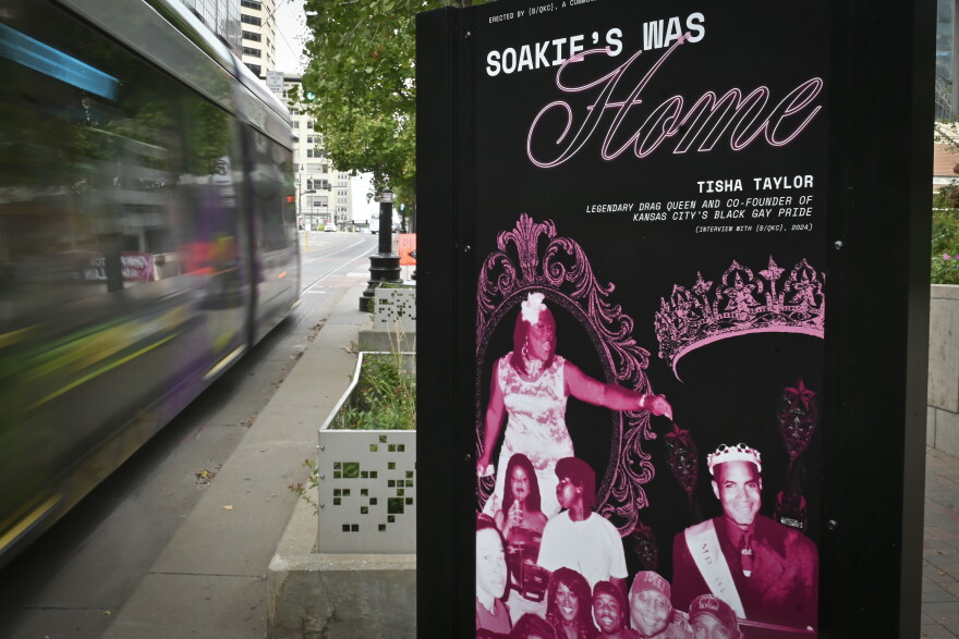 A black and purple sign sits on a city sidewalk. There are several photos of people on it. The sign reads "Soakie's Was Home."