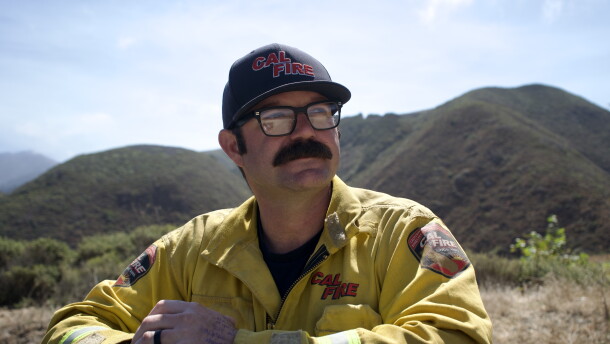 A man in a firefighting jacket with a mustache looks off into the distance with a mountain behind him. 