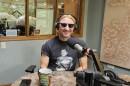 A person sitting in a radio studio with a microphone near their mouth. They have on black sunglasses.