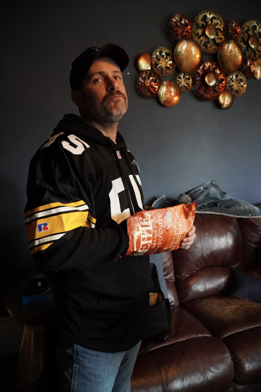 A man in a black sports jersey stands indoors holding a bag of chips. He has a neutral expression, a cap, and is in front of a brown leather couch and decorative wall art featuring metallic circular shapes.