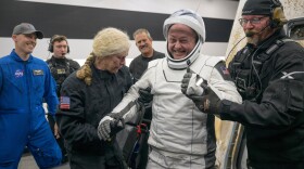 Man in white spacesuit and helmet smiles as he's helped by two people in dark, warm weather uniforms. Other people stand in the background.