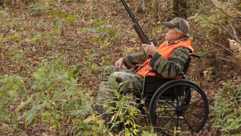 A hunter using a folding-frame wheelchair sits in a wooded area during fall. He is holding a shotgun and wearing camouflage clothing with a bright orange safety vest and camouflage cap.