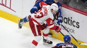 A hockey player in red and white uniform pins another player in blue uniform against the wall of an ice rink with hockey sticks in hand while another player in blue uniform comes toward them.
