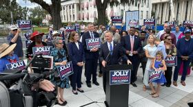 David Jolly speaks at a podium outside the Florida Capitol in Tallahassee while announcing his ticket with running mate Gwen Graham. Graham stands beside him as supporters gather around holding "Jolly Graham for Florida" signs.