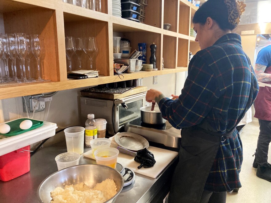 A woman in a plaid shirt and apron stirs a small pan of milk over an induction burner.