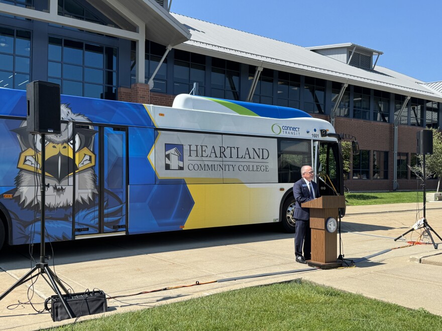A man stands speaking at a podium, with a large bus behind him. The Connect Transit bus has a hawk on it and says Heartland Community College on the windows.