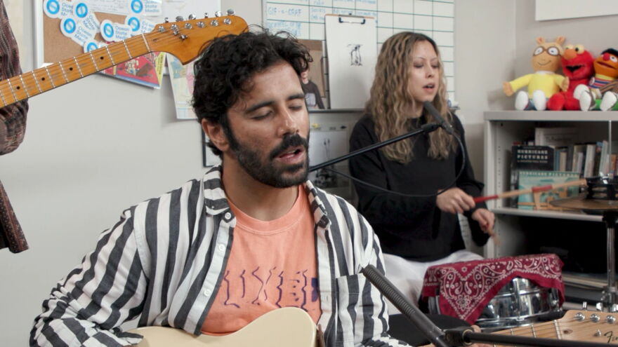 Close-up of Rafay Rashid playing guitar and singing. He's wearing a black and white striped shirt over a peach t-shirt. Talia Temporal plays a drums in the background.