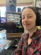 A woman wearing a floral blouse smiles in front of a microphone, various other pieces of radio equipment in the background