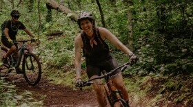 Two women ride mountain bikes on a wooded rustic trail.